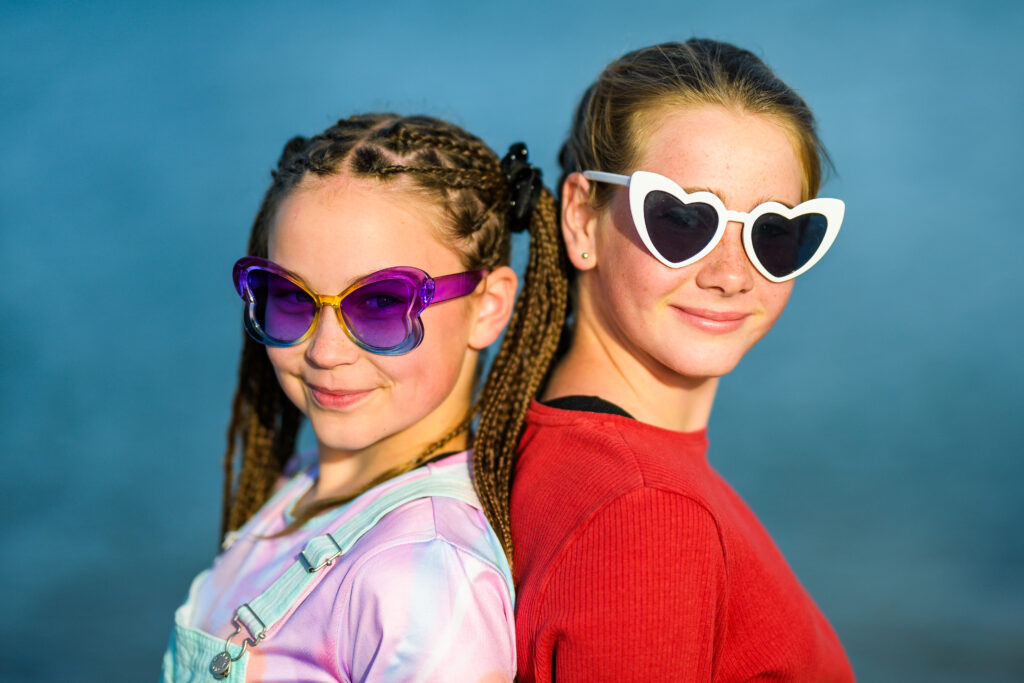 2 young people standing back-to-back, looking towards the camera. One of the young people has a rainbow T-shirt and overalls, with rainbow coloured glasses, and has their hair in lots of small braids. The other young person has a red top with white love heart shaped sunglasses. They are standing against a blue-green background.
